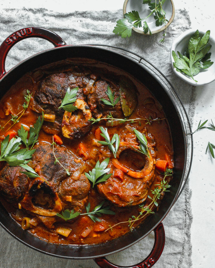 osso buco in cooking pan with bowls of herbs on a linen